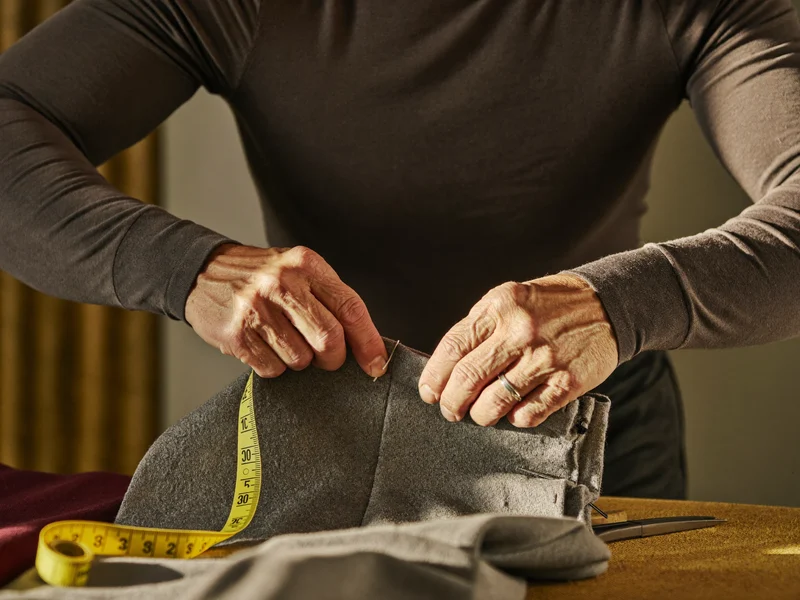 Hände stecken den Saum einer grauen Wollhose mit Kreidemarkierung und Maßband ab
